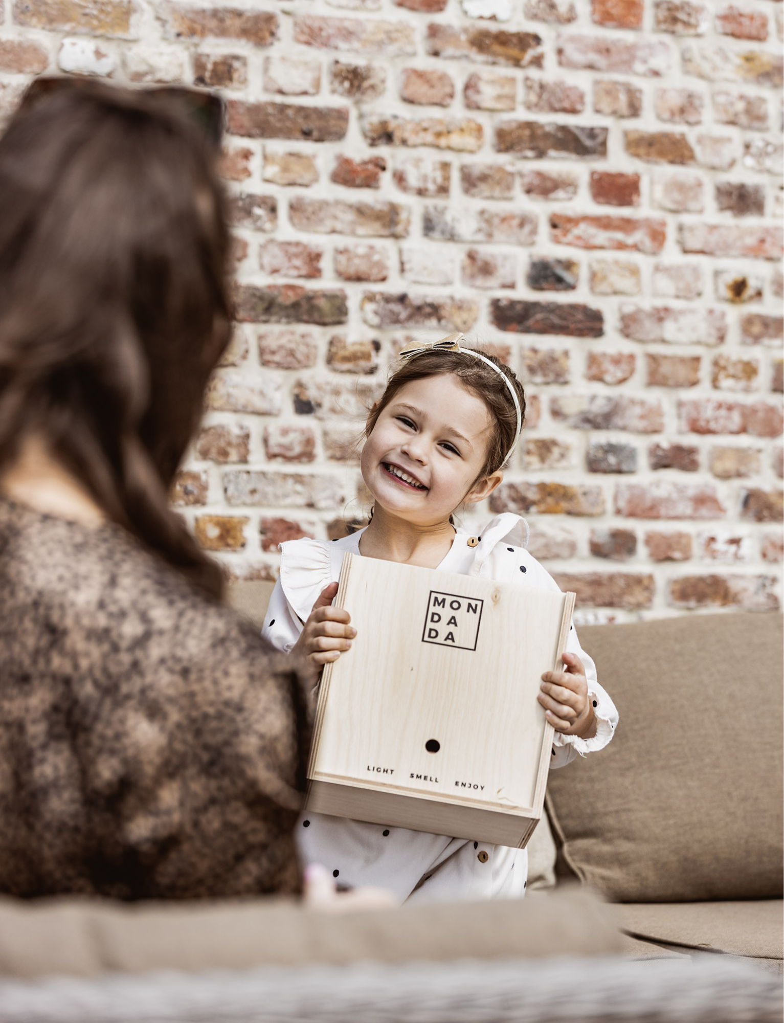 A little girl smiling and holding a Mon Dada giftbox, next to her mom.