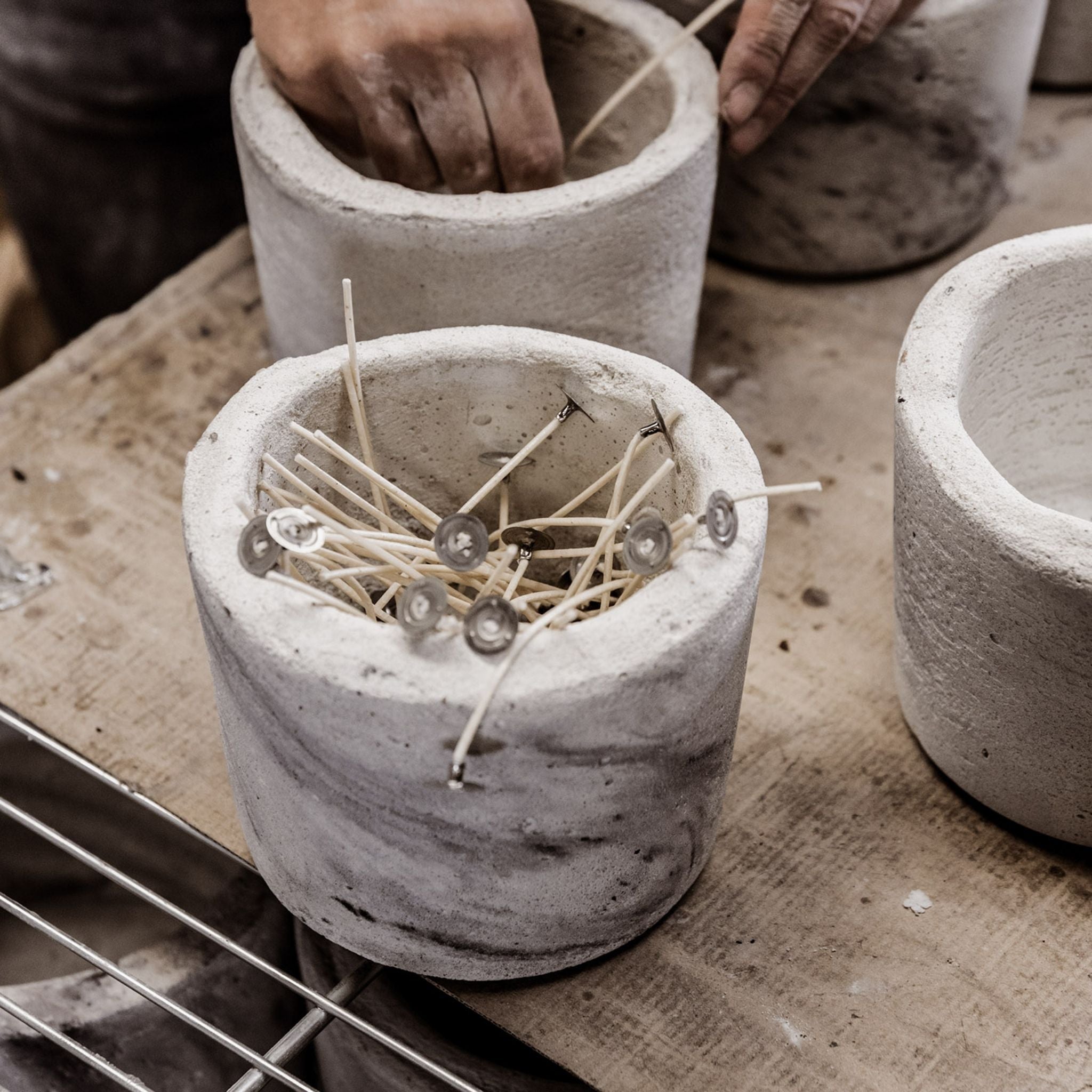 A Mon Dada candlemaker at work, crafting a handmade candle in a concrete pot. The pot sits on a wooden table in a well-lit workspace.