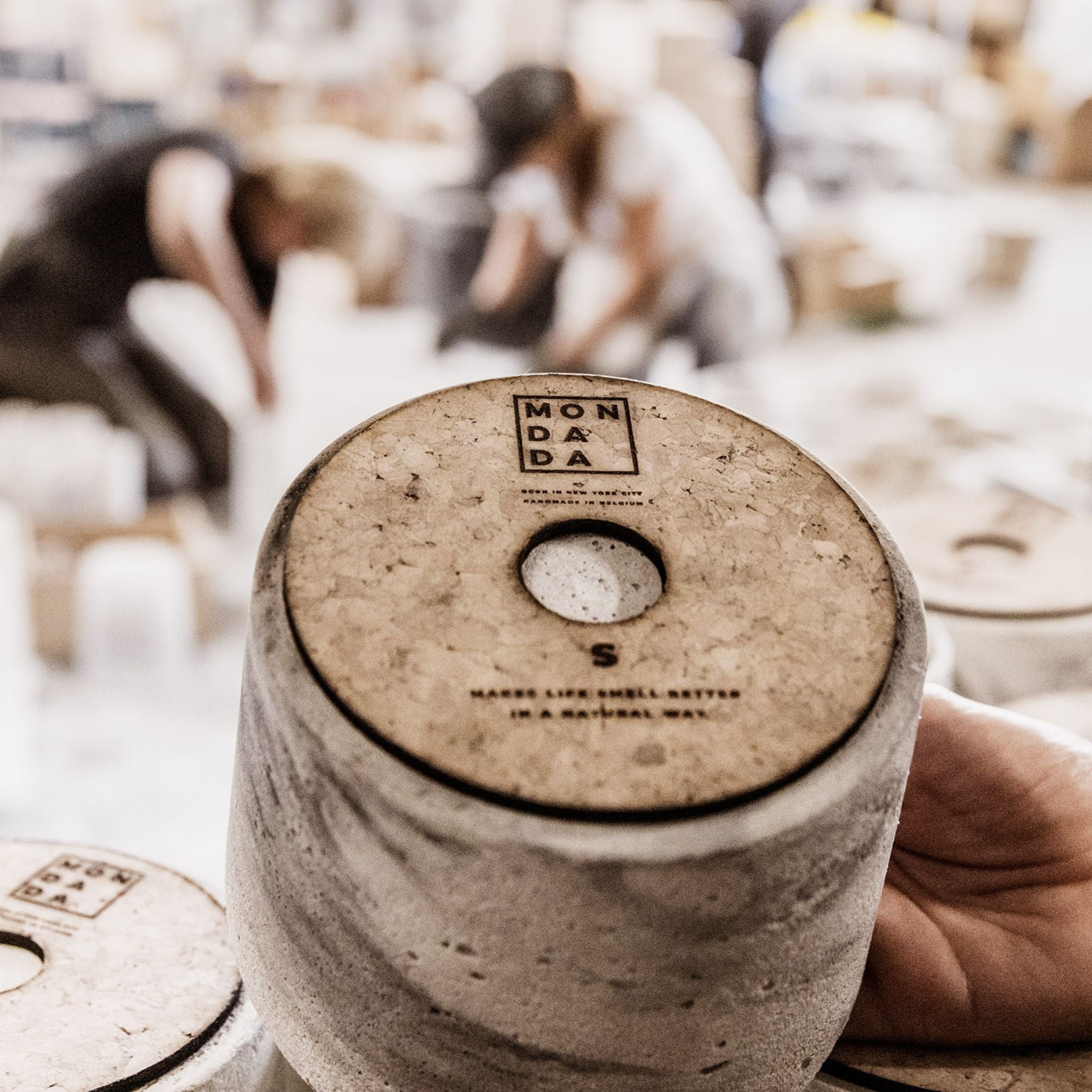 A person holding a Mon Dada candle pot. The candle is round and light grey with grey stripes in the concrete. On the bottom is a piece of cork to prevent scratches. On the cork is written "Mon Dada" and "Makes life smell better in a natural way".