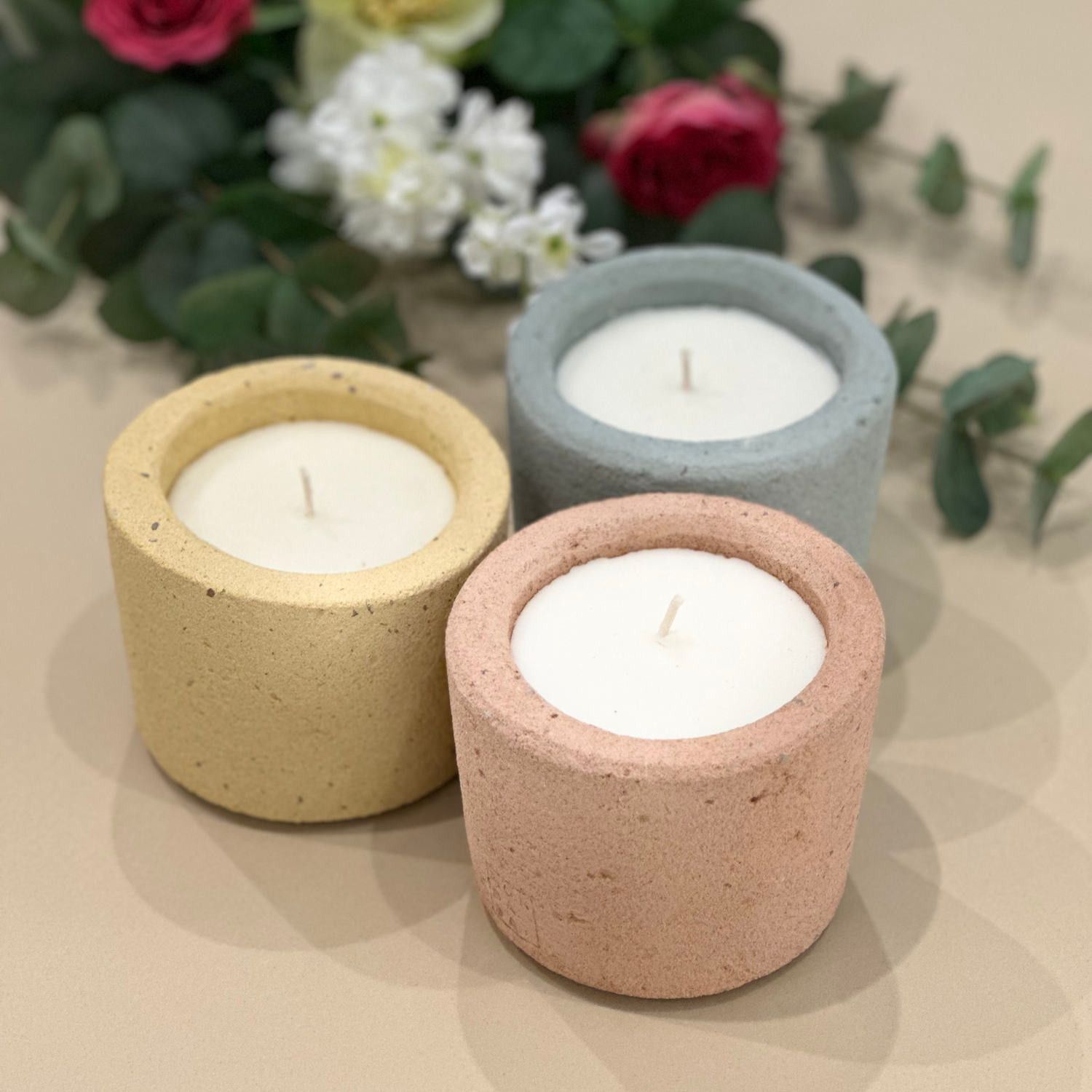 3 concrete candles in 3 colourful colours; mustard, slate and sienna. Behind them lays a bouquet of flowers.